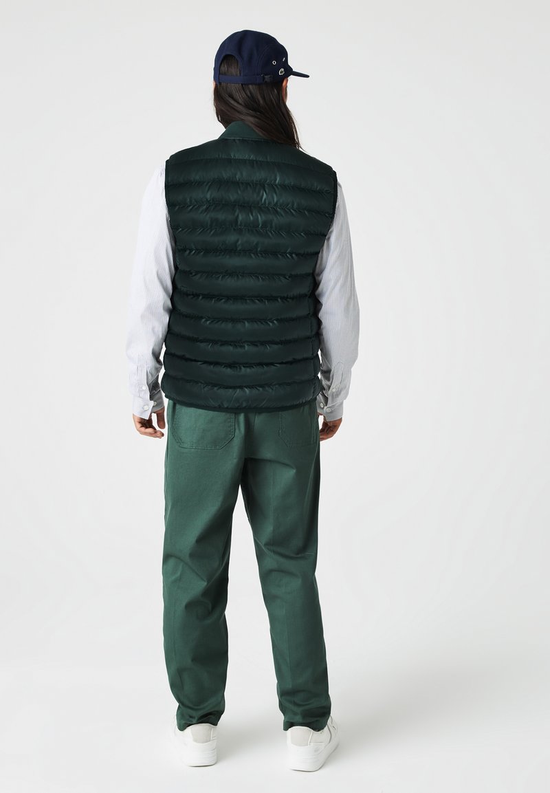 Chaleco de plumas verde oscuro con un diseño acolchado vertical, llevado sobre una camisa de botones de color azul claro. Combinado con pantalones verdes y zapatillas blancas.