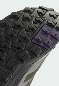 Gros plan d’une semelle de chaussure noire et violette avec des crampons profonds et robustes pour une bonne adhérence sur terrain accidenté.