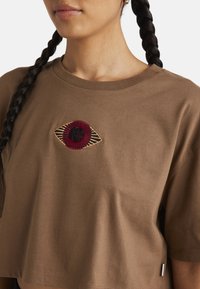 T-shirt court marron avec un design brodé aux formes d'œil rouge et noir bien visible sur la poitrine. Matière en coton doux, coupe décontractée.
