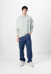 Helles grünes Hoodie mit einer Fronttasche und kleinem Logo, kombiniert mit lockeren blauen Jeans. Model trägt weiße Sneakers. Schlichter Hintergrund.