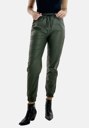 Olivgrüne Jogginghosen aus glattem, glänzendem Material, mit einem elastischen Bund und Knöchelbündchen. Kombiniert mit schwarzen Absatzstiefeln.