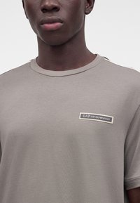 Млад човек, носещ къс ръкав EA7 Emporio Armani в цвят тауп с лого на гърдите, видим от раменете до устните.