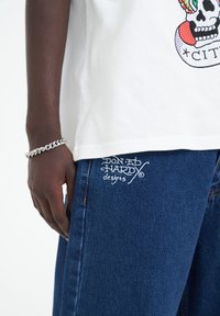 Bílý bavlněný tričko s barevným potiskem lebky, doplněné modrými džíny s bílou vyšitou značkou "Don Ed Hardy" poblíž kapsy.