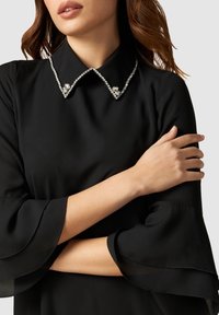 Schwarzes Kleid mit einem Kragen, der mit klaren Edelsteinen verziert ist, mit ausgestellten Ärmeln und einer glatten Textur. Mit verschränkten Armen posiert.