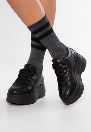 Nogi w szarych prążkowanych skarpetkach z dwoma czarnymi paskami, zestawione z masywnymi czarnymi trampkami na platformie na białym tle podłogi.