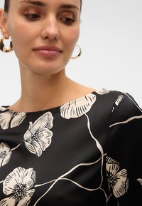 Mujer con pendientes de aro dorados y una blusa negra con grandes estampados florales blancos, mirando hacia abajo y ligeramente de lado.