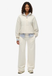 Felpa color crema con colletto zip, abbinata a pantaloni a gamba larga, indossata sopra una camicia a righe blu. Tessuto morbido e strutturato.