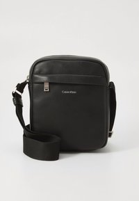 Melna Calvin Klein plecu soma ar fakturētu ādu, priekšā rāvējslēdzēja kabata un regulējamu auduma plecu siksnu uz vienveidīga fona.