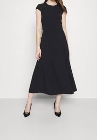 Robe noire à manches courtes avec un corsage ajusté et une jupe midi fluide. Le tissu lisse et le design simple renforcent son élégance. Talons aiguilles noirs avec des accents dorés.