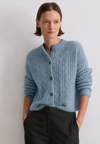 Hellblauer Strickcardigan aus weichem Material, mit Rundhalsausschnitt, fünf Knöpfen und aufgekrempelten Ärmeln. Kombiniert mit dunkler Hose.