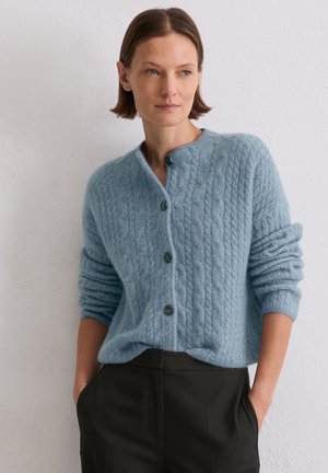 Light blue cable-knit cardigan made of soft material, featuring a round neck, five buttons, and rolled sleeves. Paired with dark trousers.
