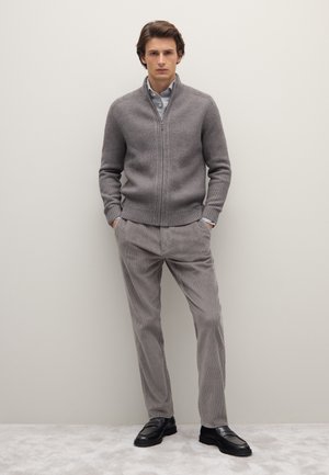 Grijze geribbelde ritsensweater over een lichtblauw shirt, gecombineerd met grijze geribbelde broeken en zwarte instappers. Eenvoudig, strak ontwerp.