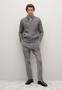 Grijze geribbelde ritsensweater over een lichtblauw shirt, gecombineerd met grijze geribbelde broeken en zwarte instappers. Eenvoudig, strak ontwerp.