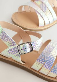 Sandali con cinturini in materiale sintetico iridescente e squamato, soletta beige e fibbia argentata. Suola piatta in gomma resistente, con un sottile motivo testurizzato.