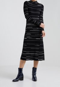 Abito nero a coste con strisce orizzontali bianche, maniche lunghe e gonna di lunghezza media. Stivaletti neri lucidi con tacco a blocco.