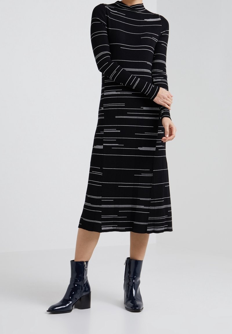 Vestido negro acanalado con rayas horizontales blancas, mangas largas y una falda de longitud media. Botines negros brillantes con tacón cuadrado.