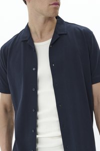 Chemise courte à manches courtes bleu marine avec un col camp, un devant boutonné et une texture lisse, portée par-dessus un débardeur blanc à côtes.