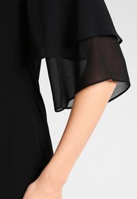 Mustapuku, jossa on liehuvat hihat läpikuultavasta kankaasta, joka on sileää ja muotoiltu istuvuutta. Yksityiskohtana hihasuunnittelu.