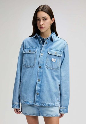 Junge Frau trägt eine übergroße hellblaue Jeansjacke und einen dazu passenden Minirock, steht vor einem schlichten Hintergrund.