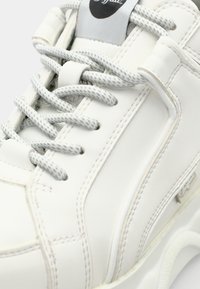 White leather sneaker with textured gray laces, featuring a rounded toe, contrasting stitching, and a logo patch on the tongue.