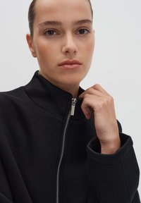 Zwarte ritssluitingjack met een hoge kraag en een gladde textuur. Zilveren metalen ritsaccent aan de voorkant. Model houdt de kraag met één hand vast.