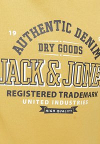 Maglietta in cotone gialla con una grafica audace in bianco e nero che riporta le scritte "Jack & Jones", "Denim Autentico" e "Alta Qualità".