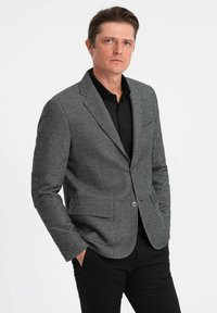 Blazer gris en espiga con dos bolsillos delanteros, cierre de un solo botón y solapas con muesca, combinado con una camisa negra y pantalones oscuros.