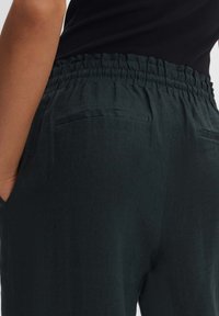 Donkergroene linnen broek met een elastische tailleband, voorzien van twee kleine zakken aan de achterkant en een gladde, gestructureerde stof.