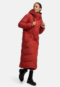 Manteau rembourré rouge avec capuche, de longueur longue, texture matelassée, deux poches latérales et accents de fermeture éclair noirs. Porté sur des bottes noires à chevilles.