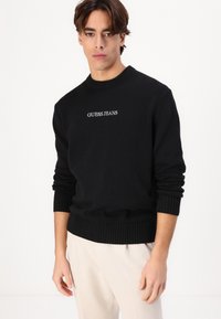 Fekete kötött pulóver bordázott mandzsettákkal és aljjal, elöl fehér "GUESS JEANS" logóval. Simaság jellemzi, klasszikus kerek nyakú dizájnnal.