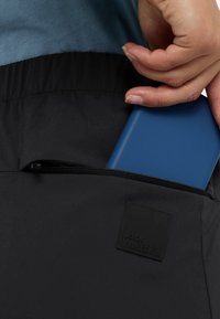 Schwarze Tasche mit Reißverschluss, in die ein blauer Artikel eingeführt wird. Sie verfügt über ein strukturiertes Logo-Patch mit erhabener Schrift.