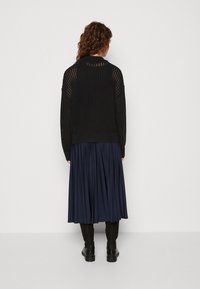 Svart stickad kavaj med öppen nätdesign, tillsammans med en plisserad marinblå kjol. Klädseln fulländas med svarta ankelboots.