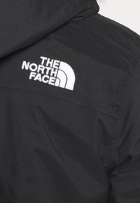 Veste noire avec un logo brodé blanc bien visible sur le côté supérieur gauche. Le tissu a une texture lisse avec un léger brillant.