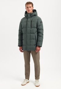 Gevulde groene jas met een hoge kraag en capuchon, voorzien van een ritszak op de borst en zwarte accenten. Gekozen met beige broeken en witte sneakers.