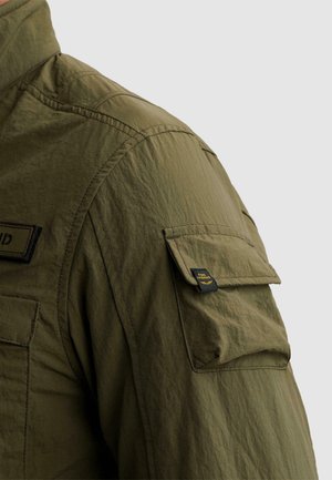 Gros plan sur la manche d'une veste vert olive avec une poche à rabat ornée d'une petite étiquette noire et jaune "PME Legend".