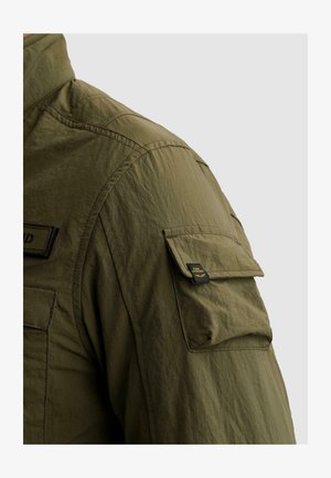 Približen posnetek rokava olivno zelene jakne z žepom s preklopom, na katerem je majhna črno rumena etiketa »PME Legend«.