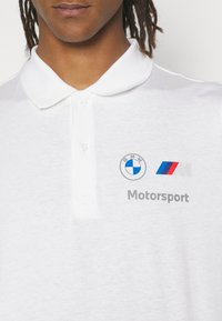 Hvid poloshirt med en blød tekstur, prydet med et blåt BMW-logo og et farverigt M-logo, samt "Motorsport" trykt i grå underneden.