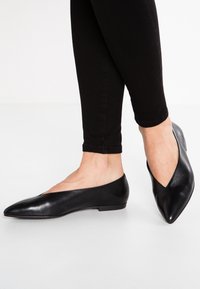 Svarta läderflatsofflor med spetsig tå och låg klack, med ett utskuret mönster längs sidorna, kombinerat med svarta åtsittande byxor.