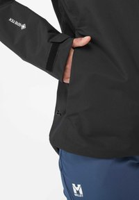 Black waterproof jacket with a zip closure and Velcro cuff. Includes side pocket and logo detailing. Worn with blue pants featuring logo.