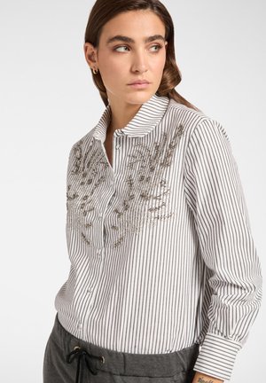 Camisa de botón a rayas en blanco y negro; presenta bordado metálico en el pecho y un cuello clásico. Se lleva con pantalones oscuros y casuales.