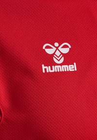 Rød mesh stof med et hvidt Hummel-logo med et bi-design over mærkenavnet, som også er i hvid skrift.