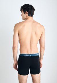 Fekete boxeralsó kék derékkal, rajta a "Lacoste" logóval, egy hátulról látható személyen, további ruházat nem látható.