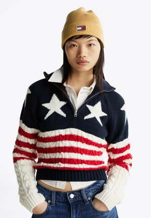 Jeune personne portant un bonnet en tricot jaune, un pull zippé avec des étoiles et des rayures, une chemise blanche à col et un jean bleu, les mains dans les poches.