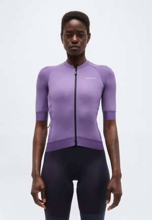 Persona atletica che indossa una maglia da ciclismo aderente a maniche corte viola con cerniera frontale e pantaloncini da ciclismo imbottiti neri, in piedi contro uno sfondo semplice.