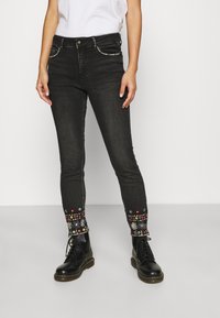 Persona vistiendo jeans ajustados negros con bordados florales coloridos cerca de los tobillos y botas negras con cordones, de pie contra un fondo blanco.