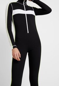 Svart långärmad jumpsuit med ett vitt horisontellt rand, framzipper och neongröna sidodetaljer. Tillverkad av stretchigt material.