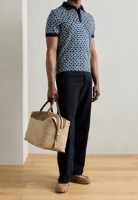 Granatowa koszulka polo z geometrycznym białym wzorem, czarnym kołnierzem i mankietami; zestawiona z czarnymi spodniami i beżową torbą podróżną ze skórzanym wykończeniem w brązowym kolorze.