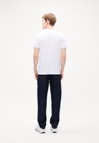 Hvit kortermet t-skjorte med rund hals, kombinert med mørkeblå denimbukser. Modellen sees fra ryggen, iført hvite joggesko.