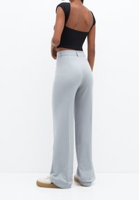 Femme portant un pantalon large bleu clair, un crop top noir à manches courtes avec un dos ouvert, et des baskets blanches avec semelles en caoutchouc, debout, vue de dos.