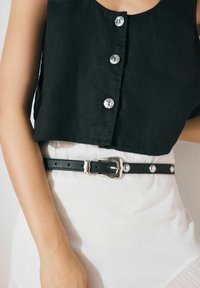 Top court en lin noir avec des boutons ronds argentés, associé à une ceinture en cuir noir avec une boucle en argent et des clous circulaires sur une jupe blanche.